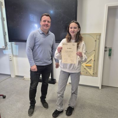 Schulleiter und neue Schülermentorin mit Urkunde vor Tafel